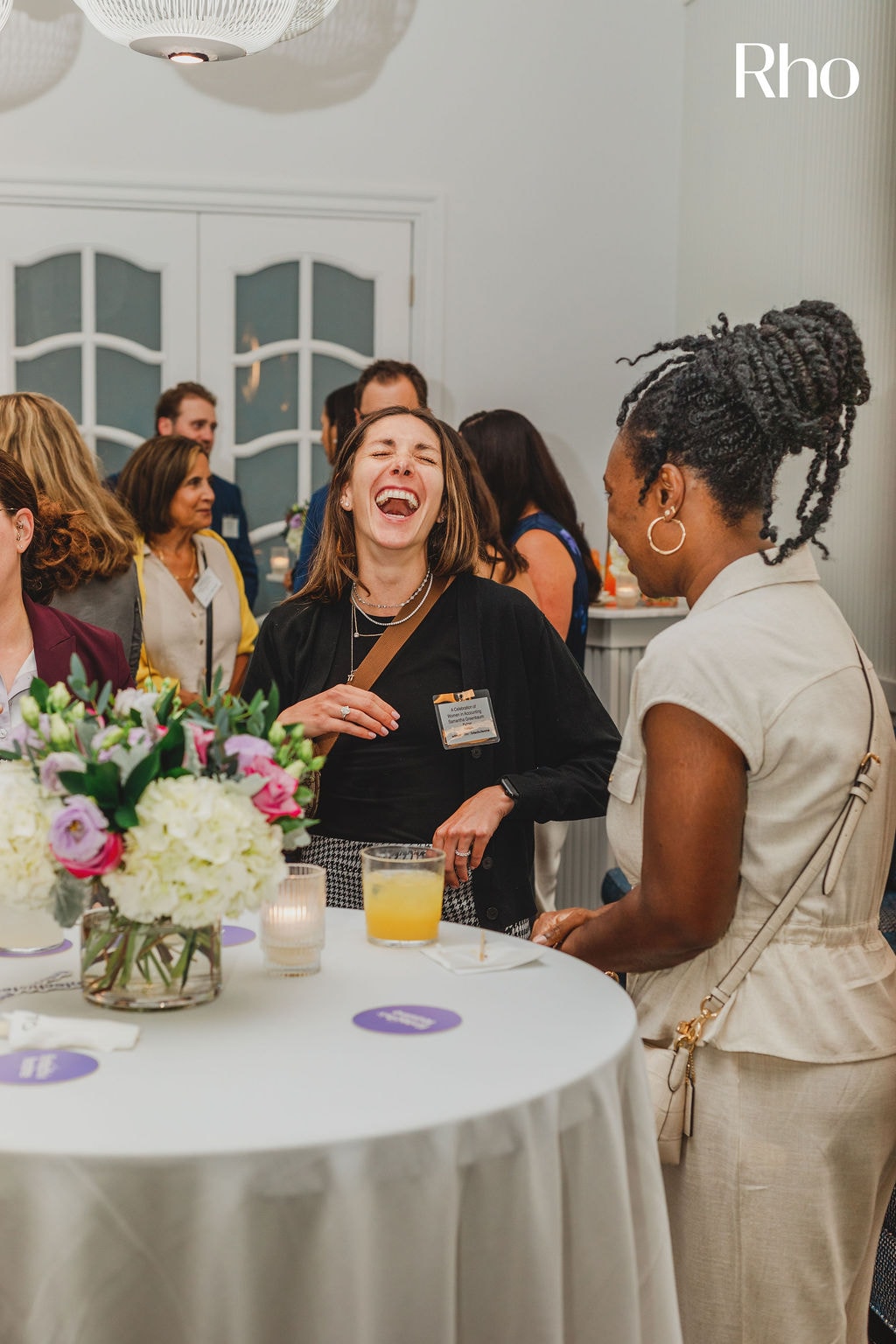 People laughing and chatting around a table with flowers and drinks at a social event.