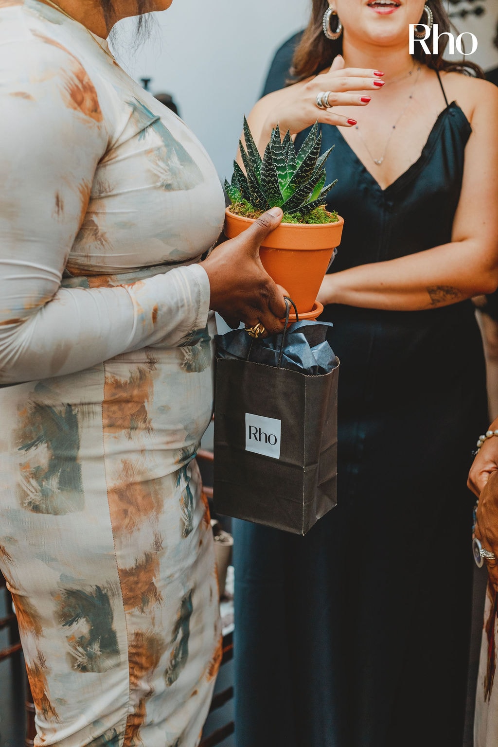 People in stylish attire holding a potted plant and a gift bag with "Rho" logo at a social gathering.