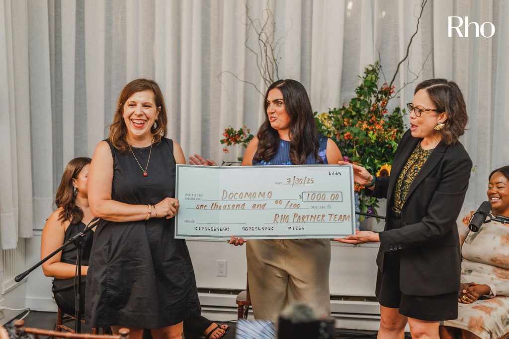 Three women stand smiling, holding a large check for $1,000, made out to "Donor Name," with a floral arrangement in the background.