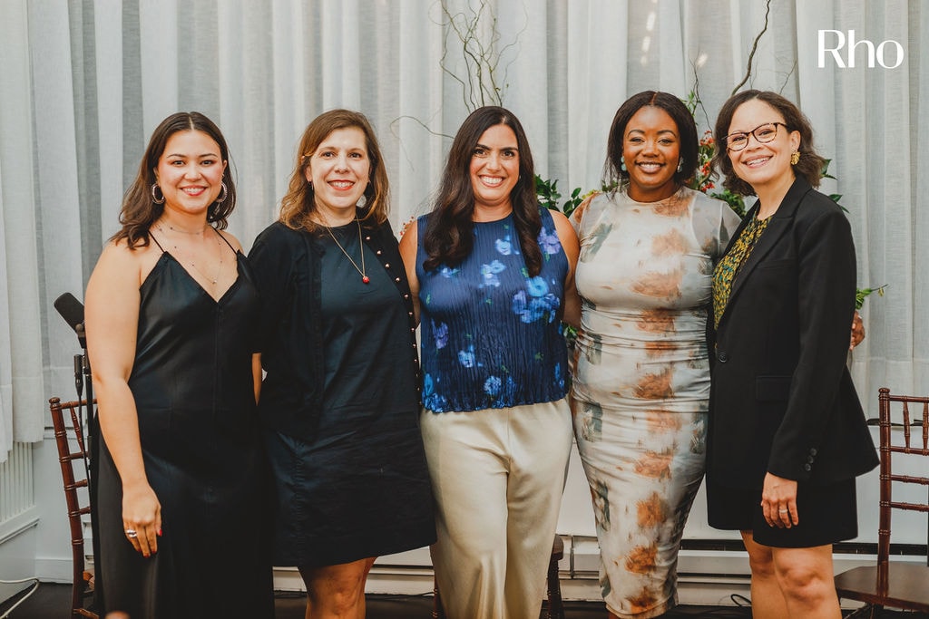 Five women standing together, smiling, in formal attire against a curtain backdrop with floral decorations.