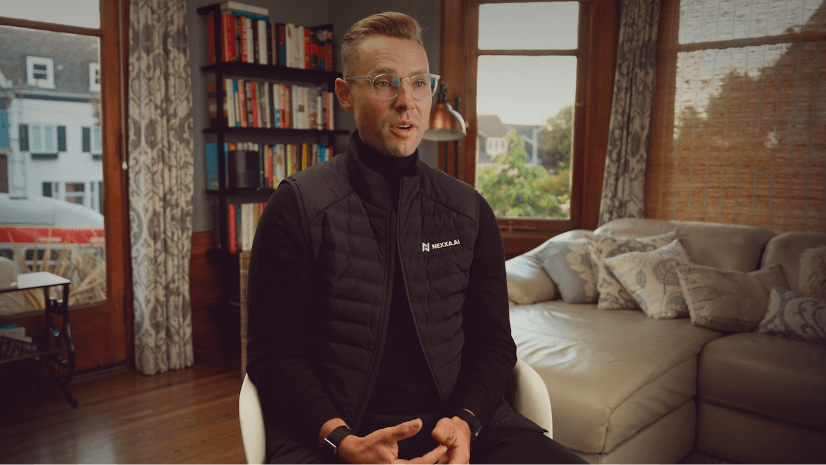 A person wearing glasses and a black jacket sits in a cozy living room with bookshelves and soft lighting.