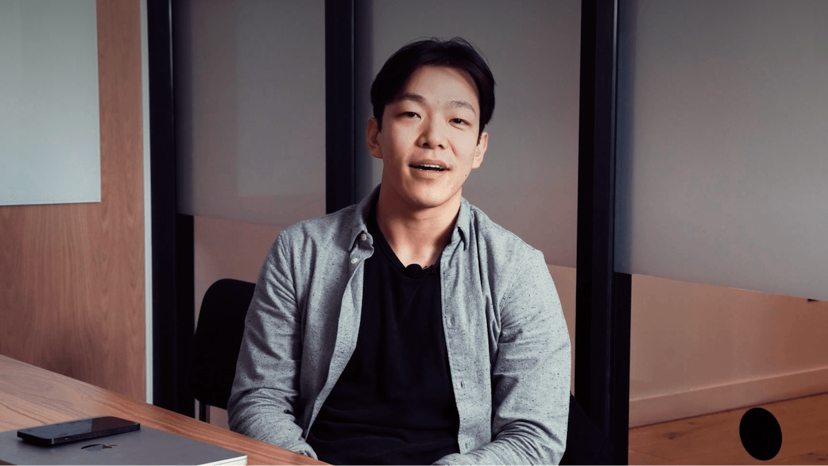 A person sitting at a desk, wearing a gray shirt over a black top, with a laptop nearby, looking towards the camera and smiling slightly.