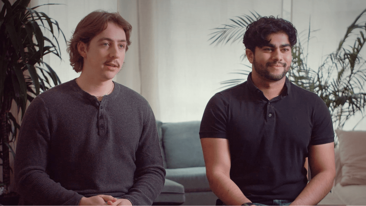 Two men sitting on a couch, facing forward. One wears a dark gray shirt; the other a black polo. They're in a room with plants and neutral decor.