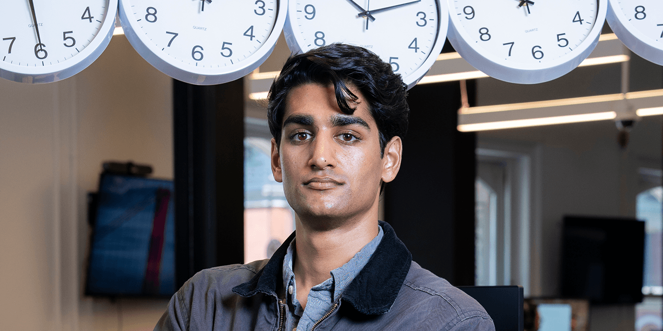 A person with dark hair stands confidently in an office, with multiple clocks on the wall behind them showing different times.