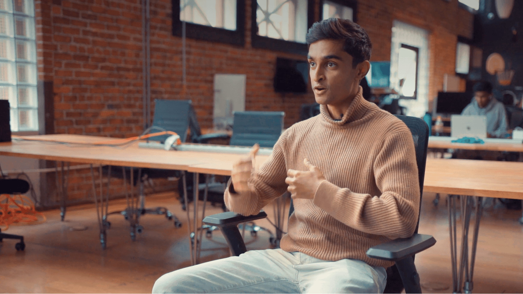 Man in tan turtleneck gesturing while seated in an open-plan office with exposed brick walls, desks and coworkers in the background.
