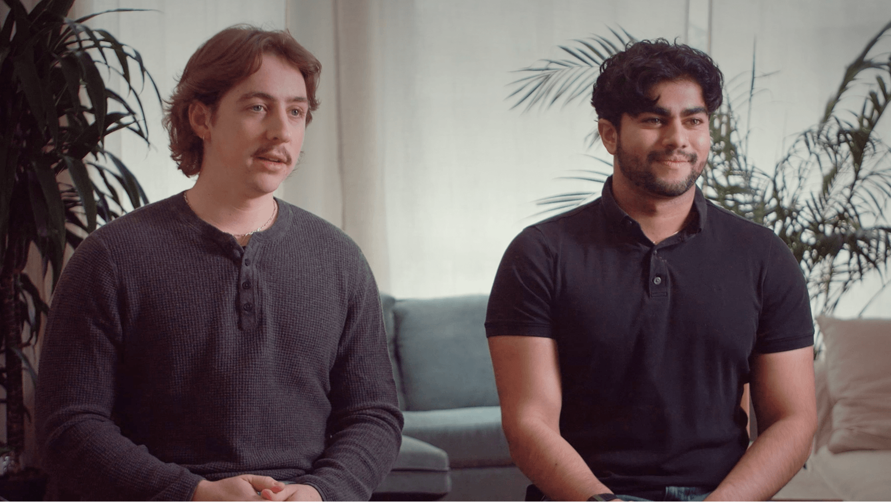 Two men seated side-by-side in a living room with potted plants behind them, one in a dark sweater and one in a black polo.
