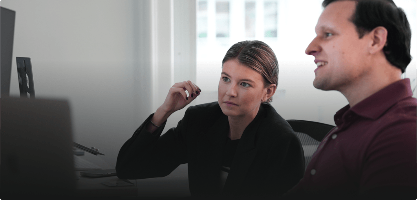 Two people sitting at a desk, engaged in a discussion while looking at a computer screen in a bright office setting.