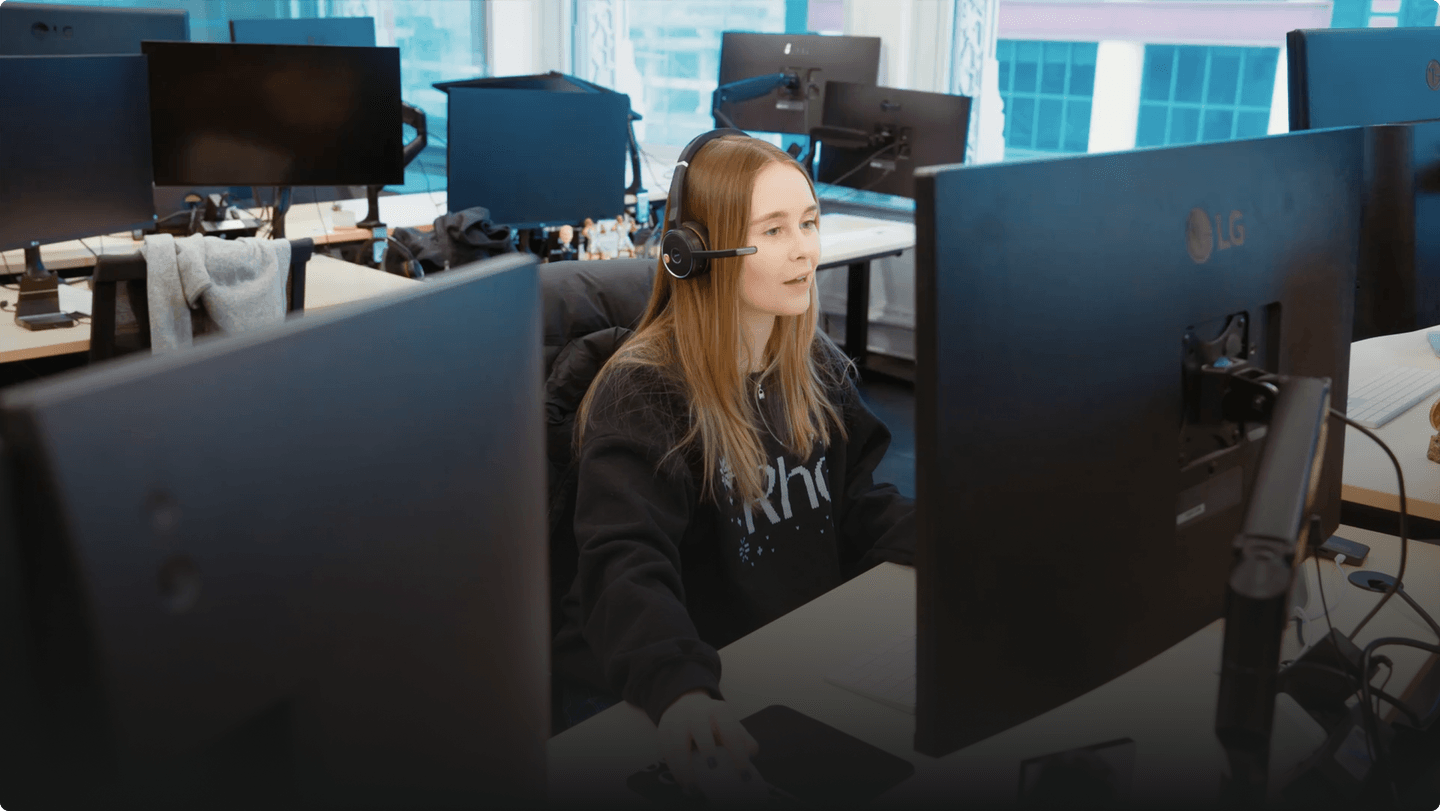 A woman wearing a headset works at a computer in a modern office with multiple monitors and large windows.