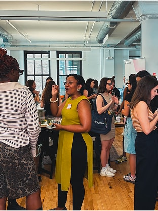 A diverse group of people socializing in a bright room with wooden floors and large windows. Some hold drinks and snacks.