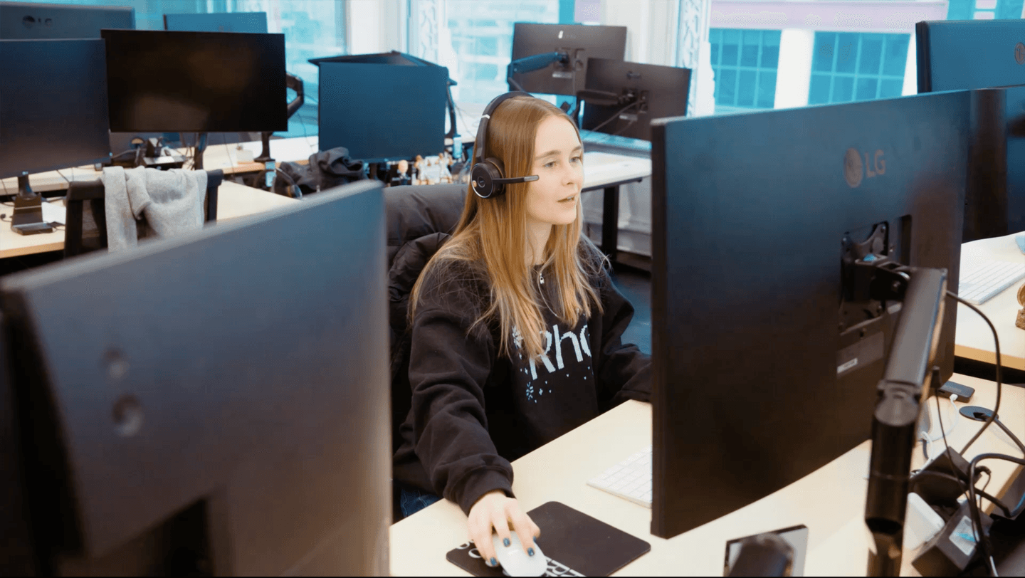 A woman wearing a headset works at a computer in a modern office with multiple monitors and large windows.