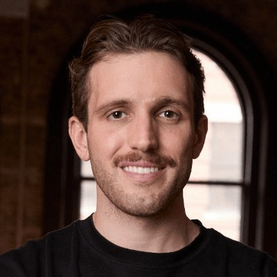 Smiling man with short hair and mustache, wearing a black shirt, stands indoors with an arched window in the background.