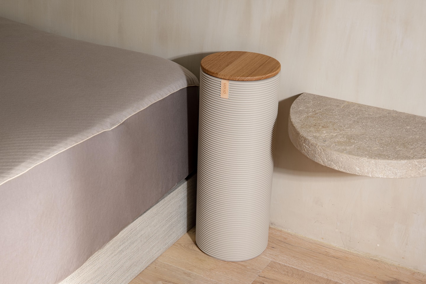A modern nightstand with a textured cylindrical design and wooden top, placed beside a bed and a curved wall shelf on a wooden floor.