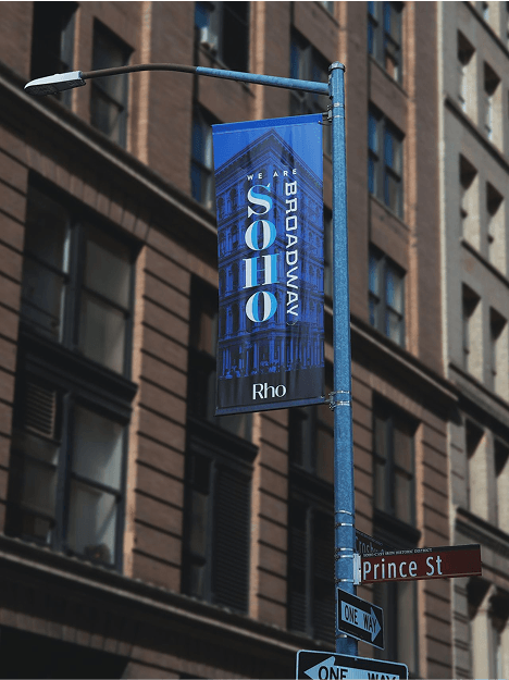 Street sign with "SOHO Broadway" banner on a pole, near a "Prince St" sign, against a backdrop of tall brick buildings.