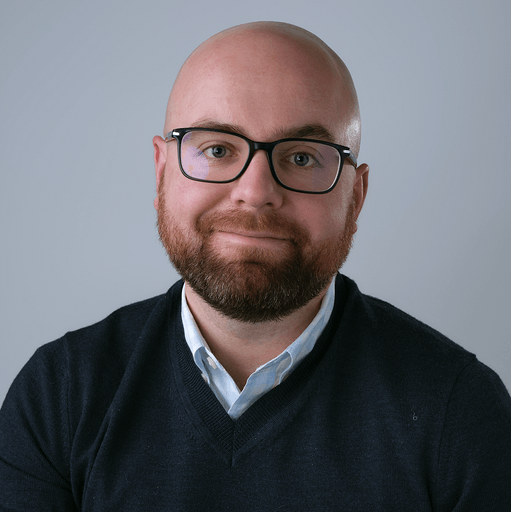 Author headshot of a man with glasses, a bald head, and a beard, wearing a dark sweater over a light collared shirt, smiles slightly against a gray background.