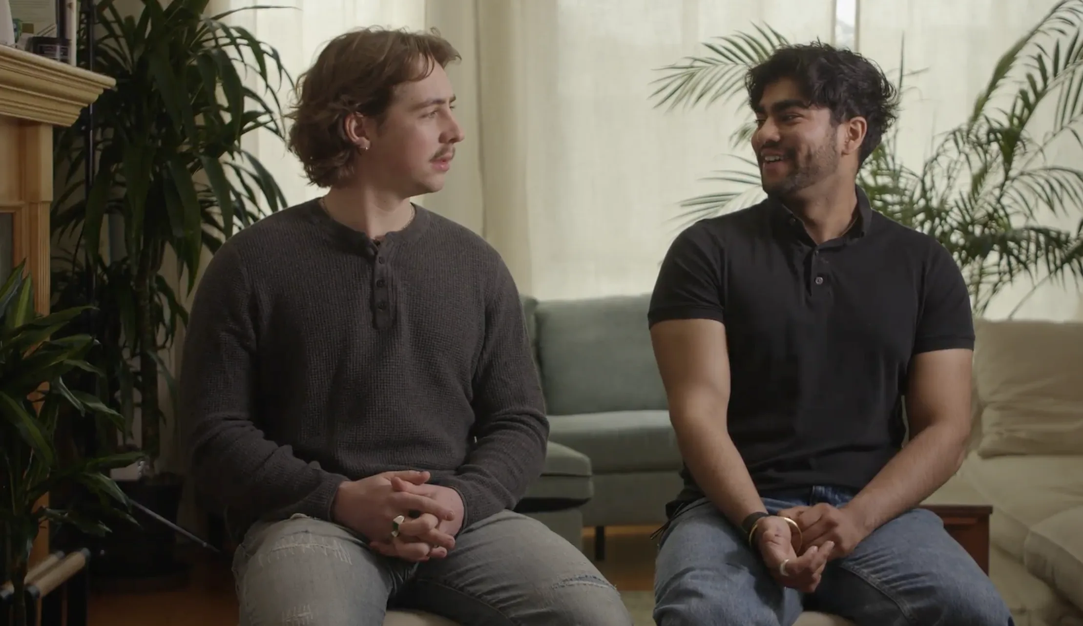 Two people seated on a couch in a living room, one turning to speak to the other.