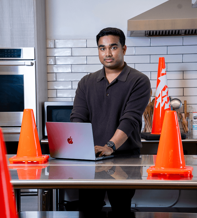 Person standing in a kitchen, working on a laptop at a counter with orange traffic cones. Oven and tiled wall in the background.