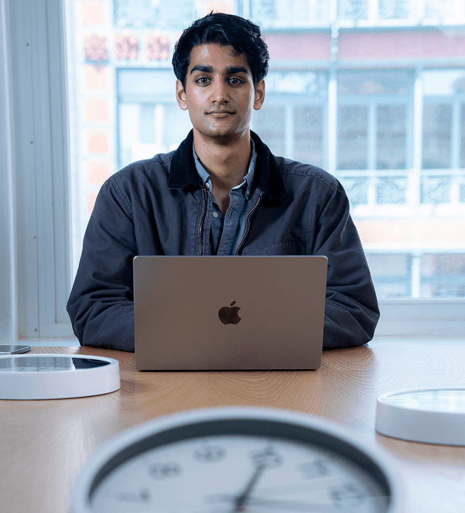 Person sitting at a table with a laptop, facing the camera. A clock is blurred in the foreground, and a large window is in the background.