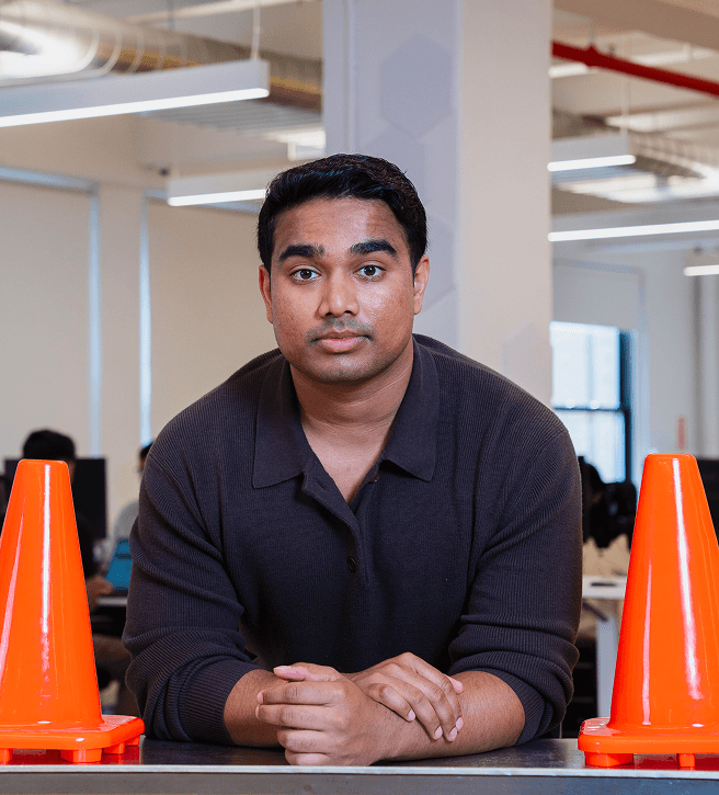 Man in a brown sweater leaning on a table with two orange traffic cones in an office setting.