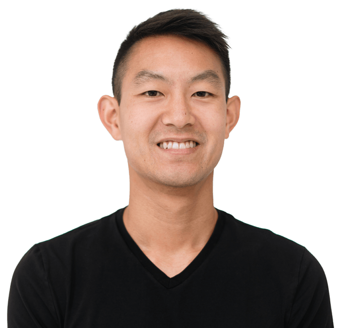 Smiling person with short hair wearing a black V-neck shirt, set against a plain white background.