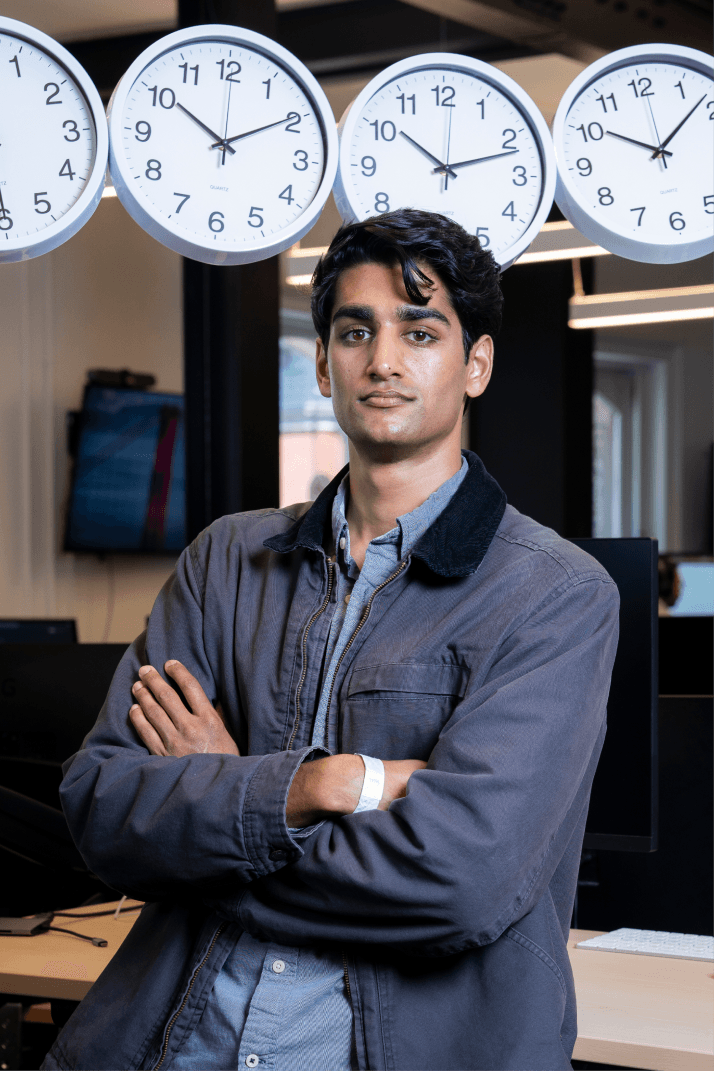 Person with crossed arms, wearing a gray jacket, stands confidently in front of five wall clocks showing different times.
