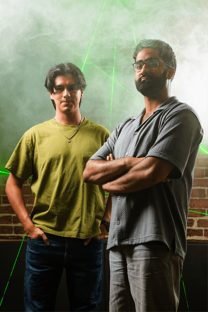 Two men stand confidently in a smoky room with green laser lights. One wears a green shirt, the other a gray shirt. Brick wall in the background.