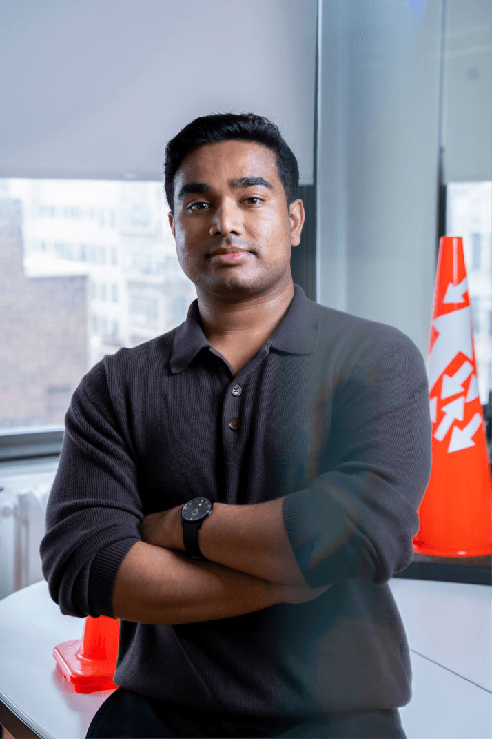 A person standing with arms crossed in an office, wearing a dark shirt. A traffic cone is visible in the background near the window.