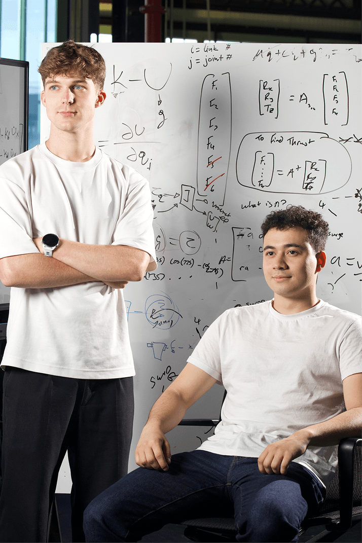 Two young men in white shirts, one standing and one seated, in front of a whiteboard filled with mathematical equations.