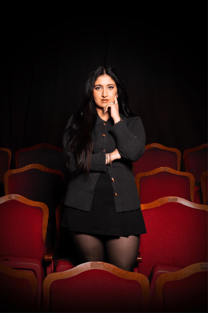 Woman standing confidently among red theater seats, wearing a dark outfit, with long dark hair. The background is dimly lit.