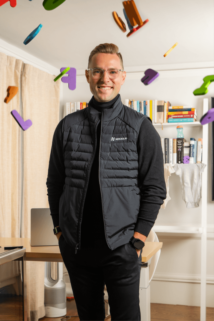 A person smiling in an office space with bookshelves, a laptop, and colorful objects suspended in the air.