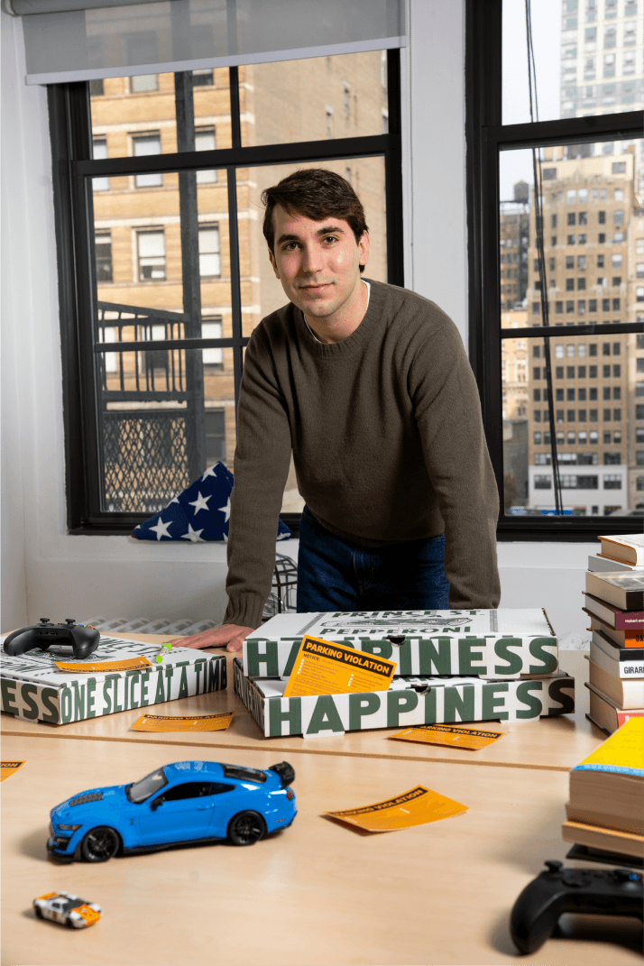 Man leaning on a table with board games, a toy car, books, and a video game controller. Urban window view in the background.