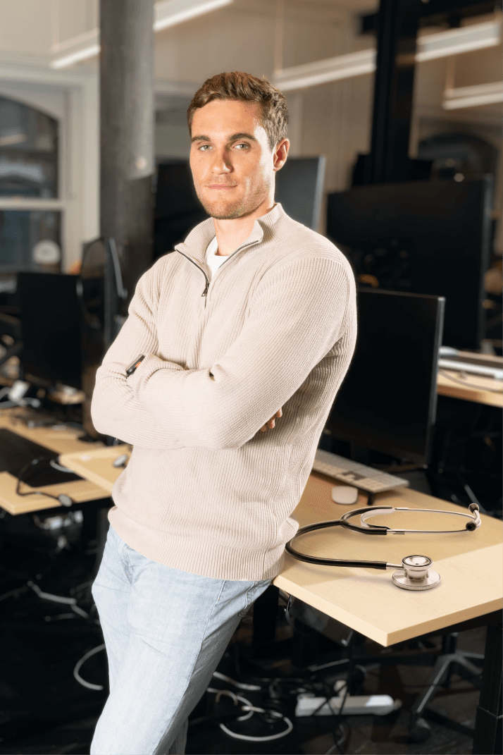 A person in a beige sweater and jeans stands with arms crossed in an office, a stethoscope on the desk nearby.