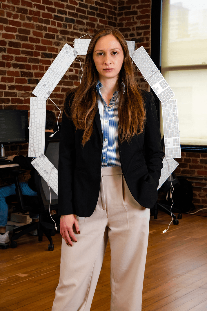 A woman in a blazer stands in an office setting with a circle of keyboards behind her, in front of a brick wall and window.