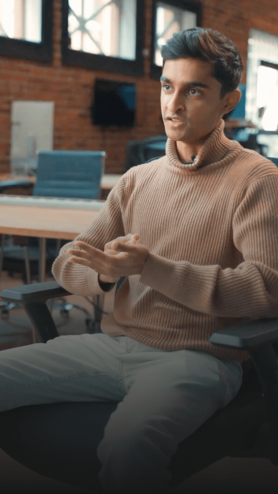 Person in a brown turtleneck sweater sits in an office chair, gesturing with hands, with a blurred office background.