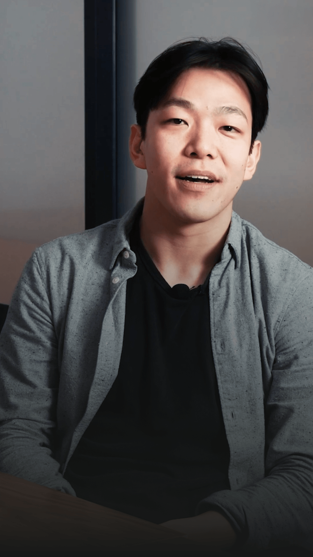 A person with short dark hair, wearing a black shirt and gray button-up, is speaking and facing forward.