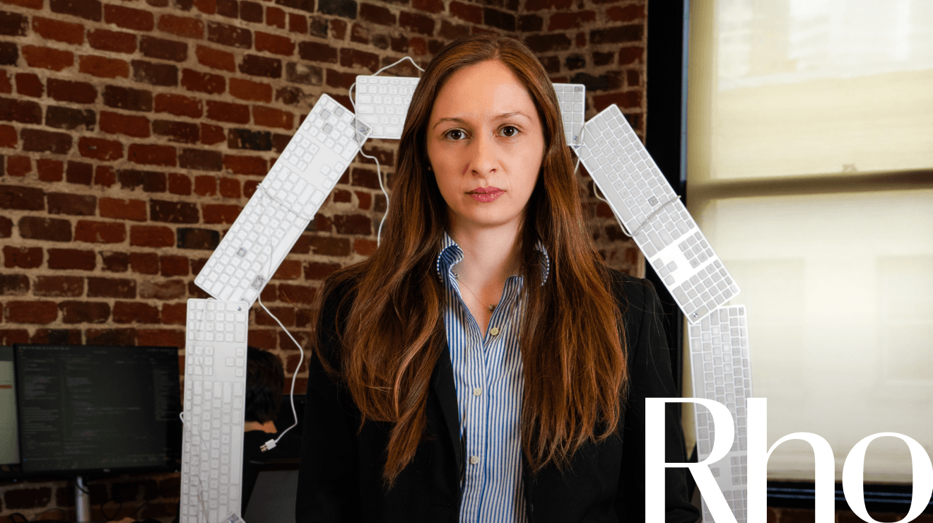 Woman in a business suit stands confidently in front of a brick wall with a keyboard arch behind her. Office setting with computer screens visible.