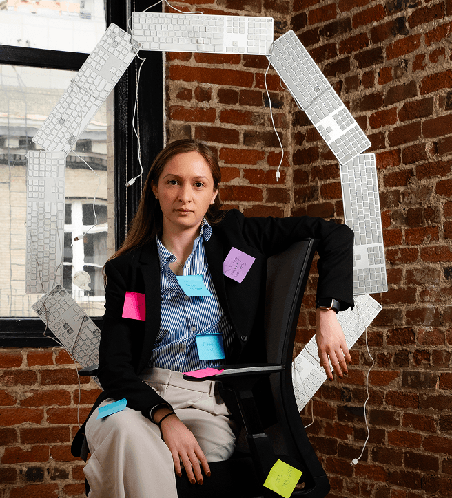 A woman in a blazer sits in an office chair, surrounded by a circle of keyboards, with sticky notes on her clothes, against a brick wall.